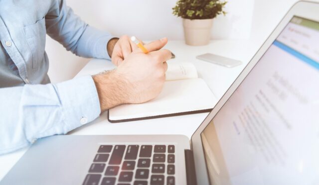A man writing in a notebook on a desk with a laptop and smartphone, in a modern office setting.