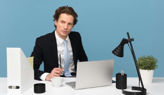 A focused businessman in a suit works at his tidy desk with a laptop, plant, and lamp.