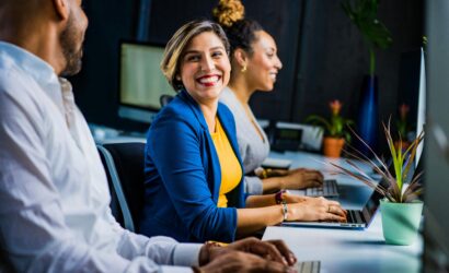 Three diverse professionals working and smiling at office desks, fostering teamwork and collaboration.