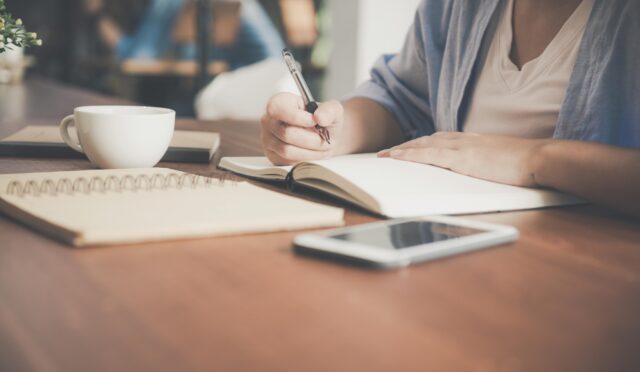 A woman writes in a notebook at a café table with a coffee and smartphone nearby.