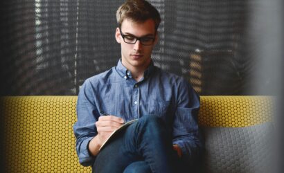 A young man in glasses writes in a notebook while sitting on a stylish couch indoors.