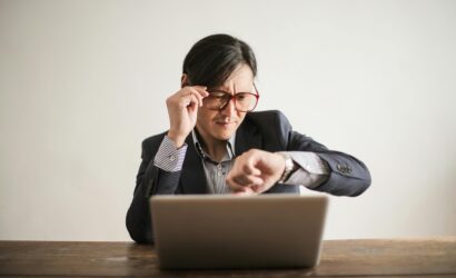 Young frowning man in suit and glasses looking at wristwatch while waiting for appointment sitting at desk with laptop