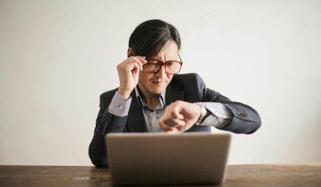 Young frowning man in suit and glasses looking at wristwatch while waiting for appointment sitting at desk with laptop