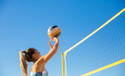 A woman plays volleyball on a sunny day, reaching to hit the ball over the net.