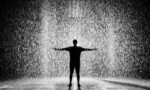 A person joyfully standing in the rain with arms wide open in a dramatic black and white scene.