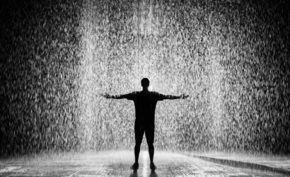 A person joyfully standing in the rain with arms wide open in a dramatic black and white scene.