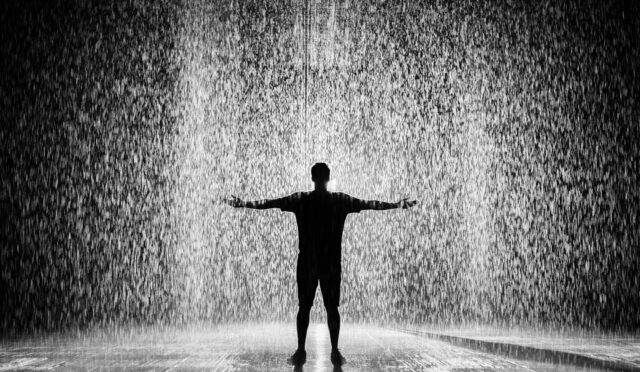 A person joyfully standing in the rain with arms wide open in a dramatic black and white scene.
