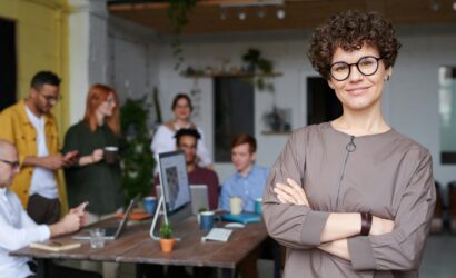 Smiling businesswoman with curly hair stands confidently in a modern office space with colleagues.