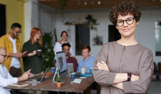 Smiling businesswoman with curly hair stands confidently in a modern office space with colleagues.