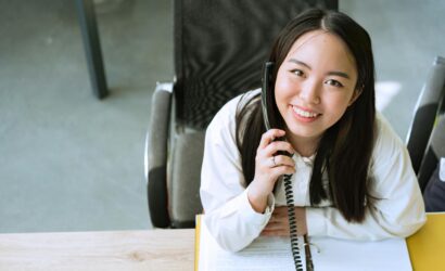 Smiling woman in a white shirt talking on the phone while sitting at an office desk.