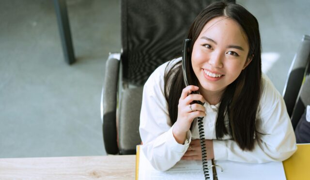 Smiling woman in a white shirt talking on the phone while sitting at an office desk.