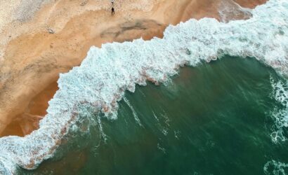 Aerial shot of ocean waves crashing onto a sandy beach with two people in the distance.