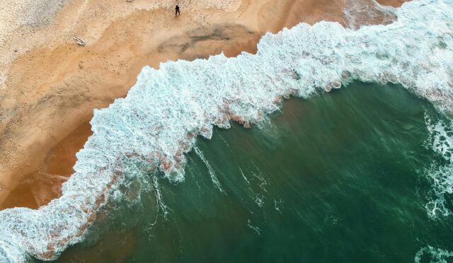 Aerial shot of ocean waves crashing onto a sandy beach with two people in the distance.