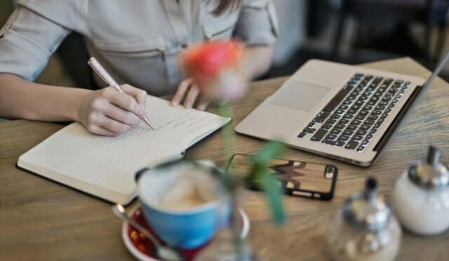 Woman writing in a notebook with a laptop and coffee cup on a desk. Ideal for workspace inspiration.