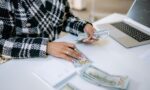 Close-up of a woman counting hundred-dollar bills at a desk with a laptop, focusing on finance and work.