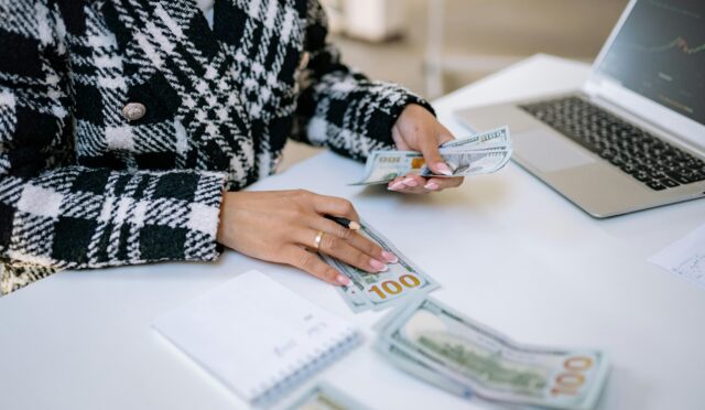 Close-up of a woman counting hundred-dollar bills at a desk with a laptop, focusing on finance and work.