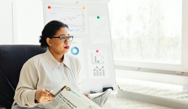 Confident businesswoman reading newspaper and analyzing market trends in a modern office setting.